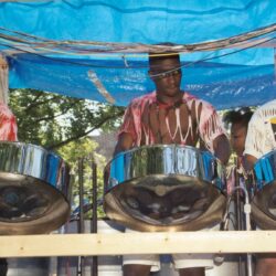 Three men playing steelpan drums under blue tarp