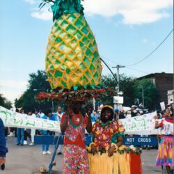 Two performers in colorful outfits carrying giant pineapple